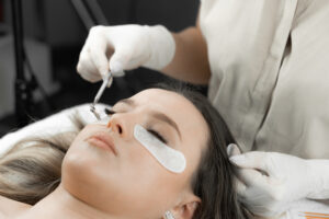 A beautician looks in a mini mirror and looks at the newly extended eyelashes of a young woman in a beauty salon. Woman’s face in cosmetic procedure close-up